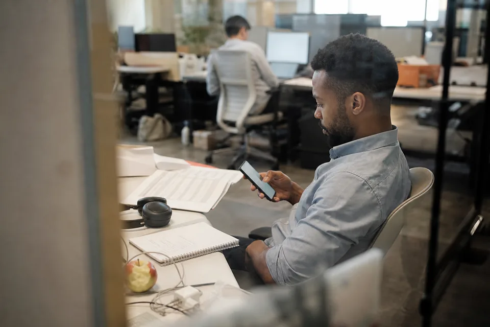 A sales lead leaning back at his desk reading his phone next to a printed proposal and a handwritten checklist