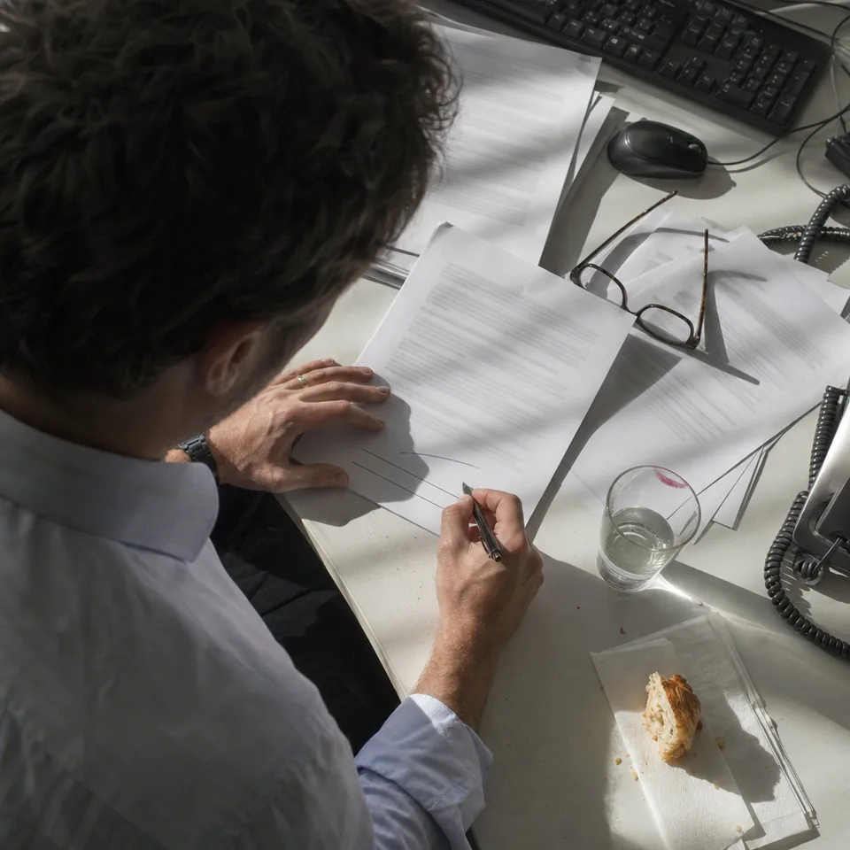 A hand with a pen mid signature on the last page of a printed proposal on a cluttered white desk, seen from above